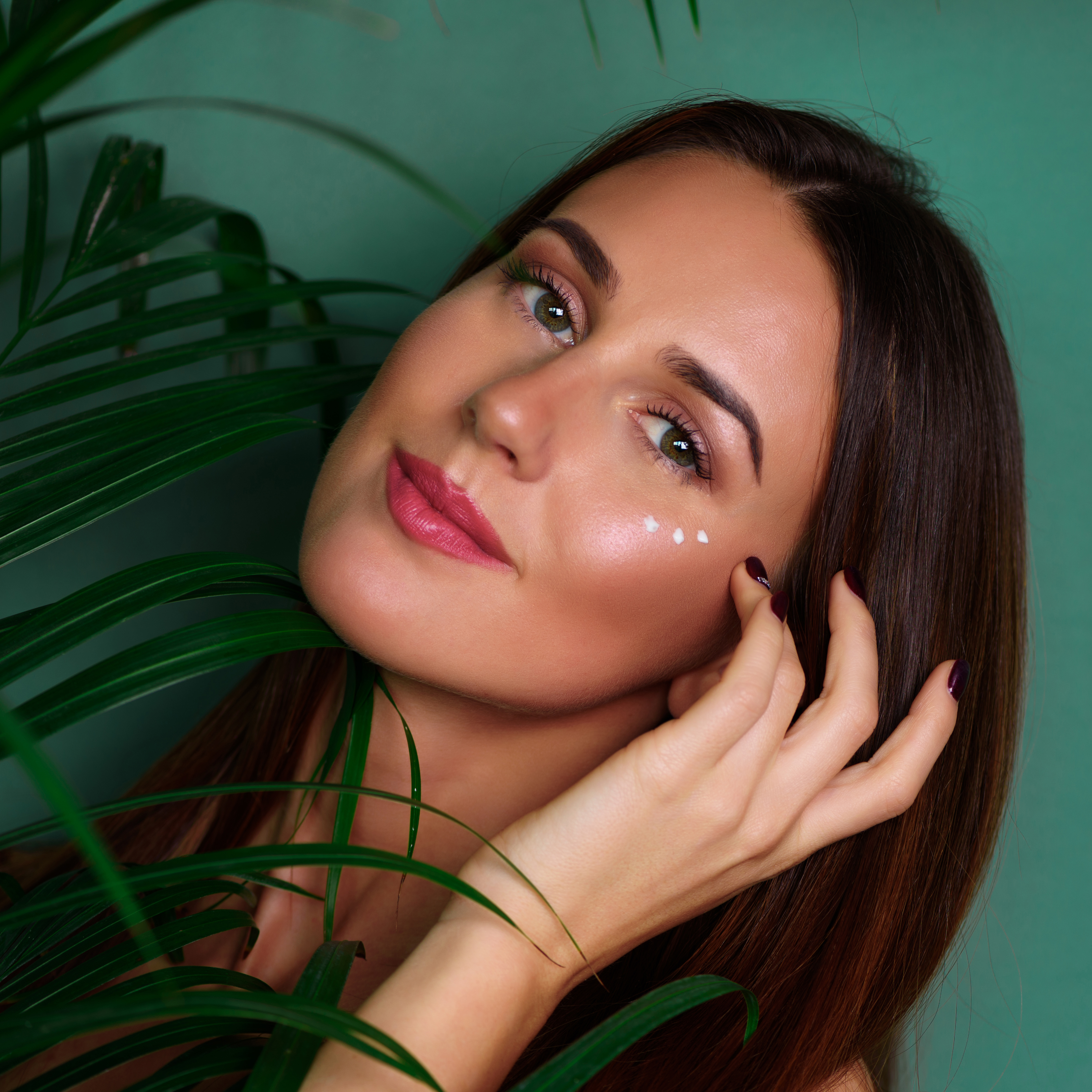 Girl with cream dots on her face between palm leaves on green background. Copy space. Beauty, healthy skin, body care, natural make up concept. Square crop